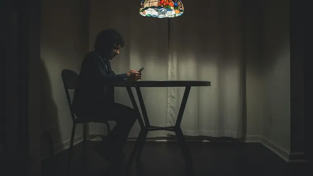 A lone figure at a table in a dim room, lit by a phone